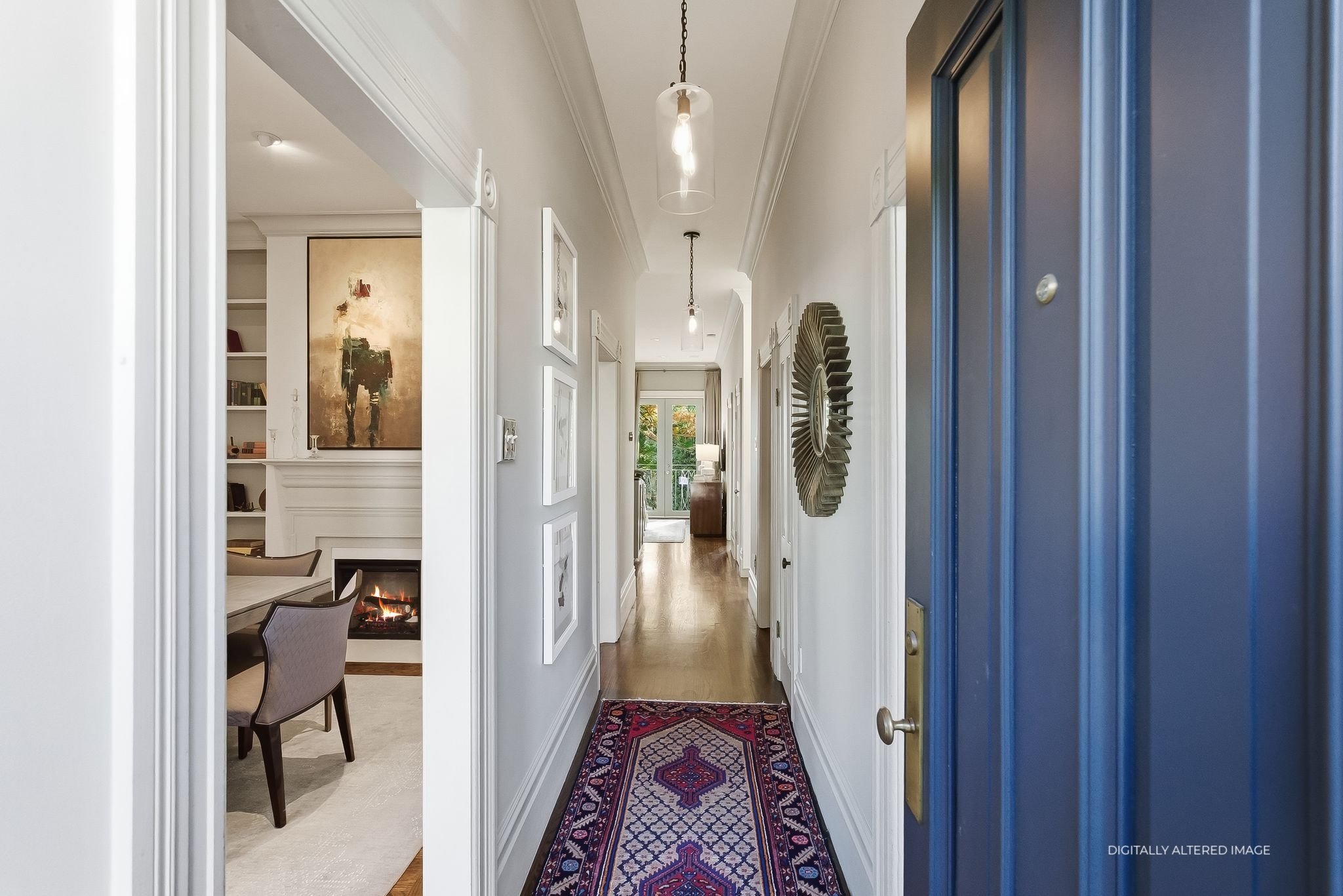Interior hallway of a house with white walls, hardwood floors, decorative wall art, and a colorful patterned rug. Visible through the doorway on the left is a room with a fireplace, chairs, and other furniture.