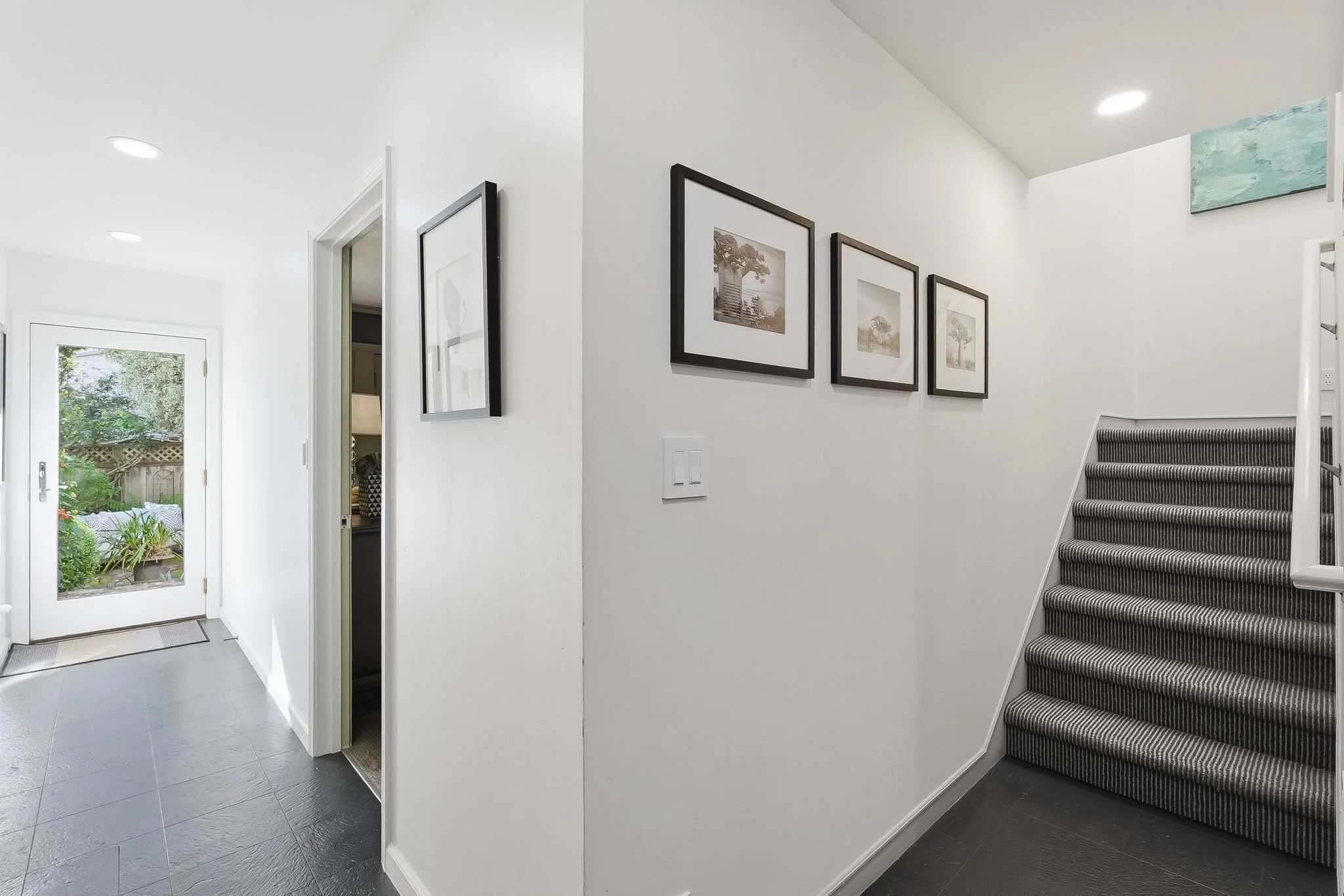 Interior view of a modern home with white walls, a staircase covered in dark striped carpet, framed artwork on the wall, a glass door leading outside, and a glimpse of a garden through the door.