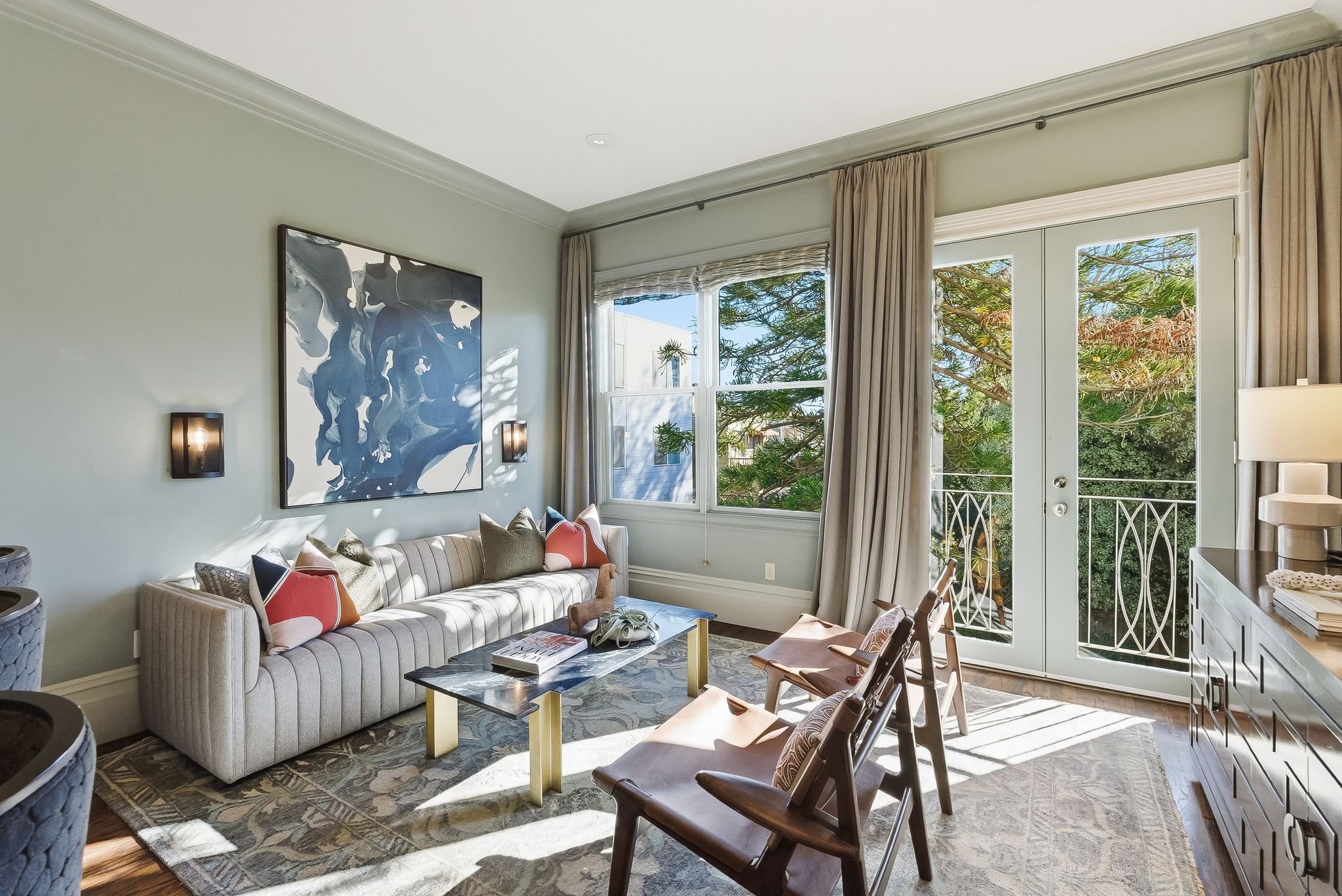 Bright living room with large windows and a glass door leading to a balcony, featuring a striped sofa with colorful pillows, a marble-top coffee table, wooden chairs, a sideboard with a lamp, and greenery outside.