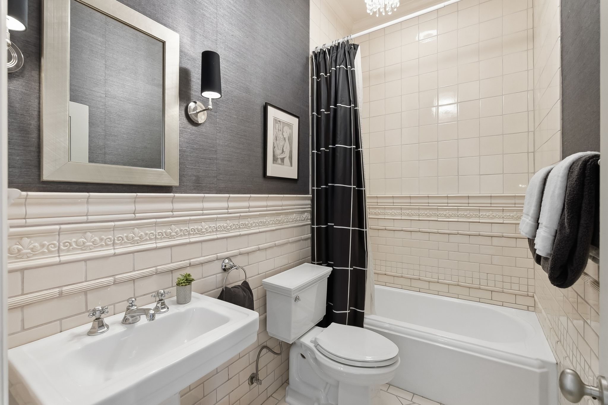 A small bathroom with a white sink, a mirror, a black wall sconce, a framed picture, a white toilet, and a bathtub with a black and white shower curtain. There are towels hanging on a rack.