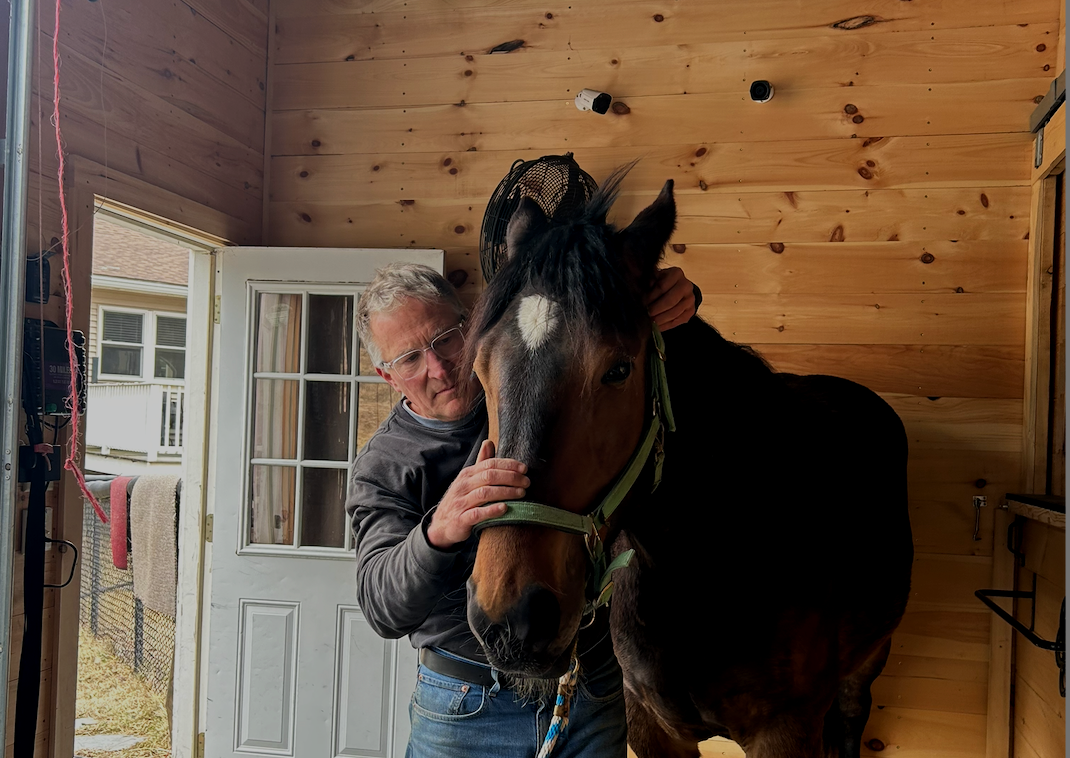 An older man with glasses gently petting a brown and black horse inside a wooden stable.