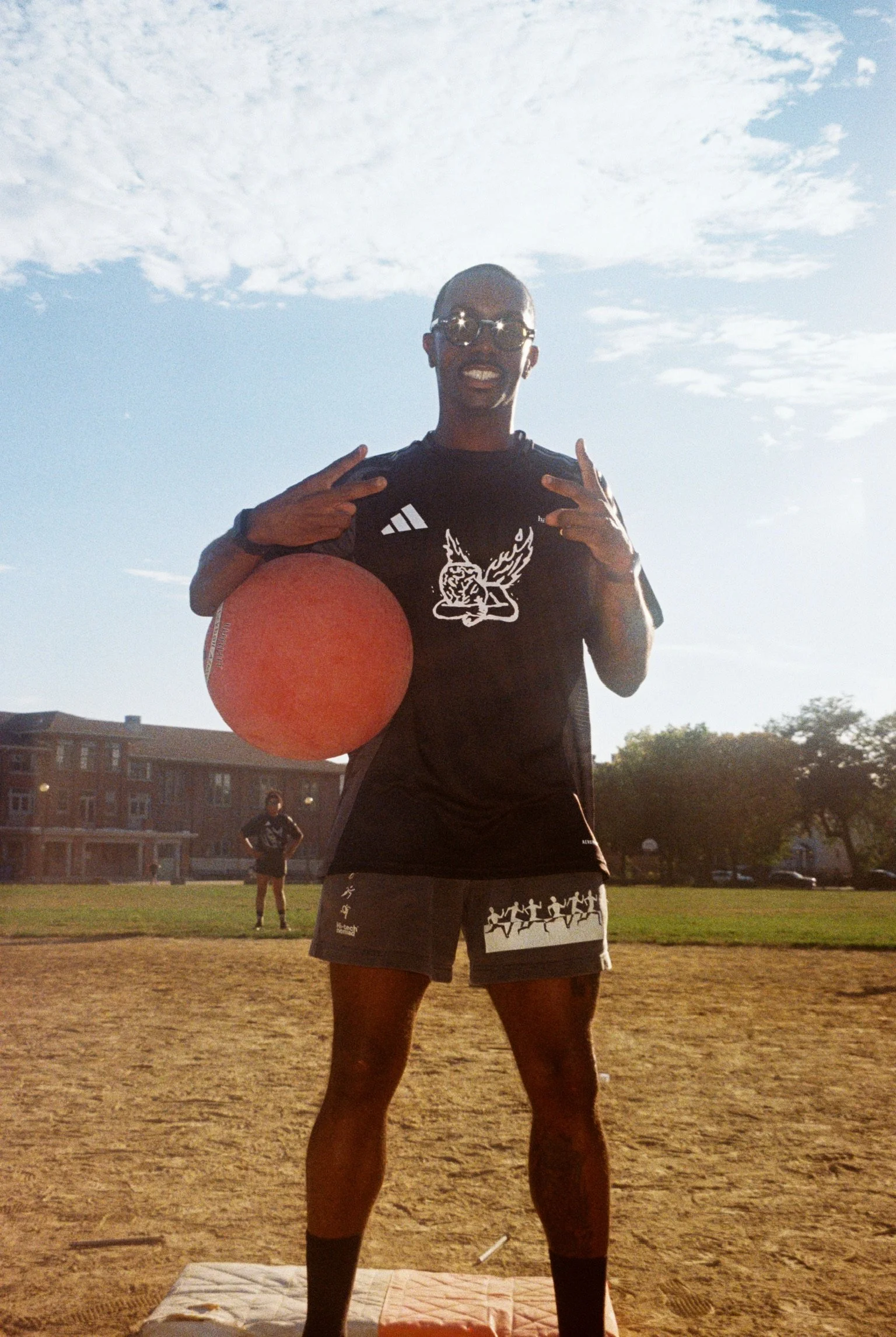 A person standing outdoors on a sandy field, holding a red volleyball, wearing sunglasses and sportswear, smiling at the camera, with sunny sky and buildings in the background.