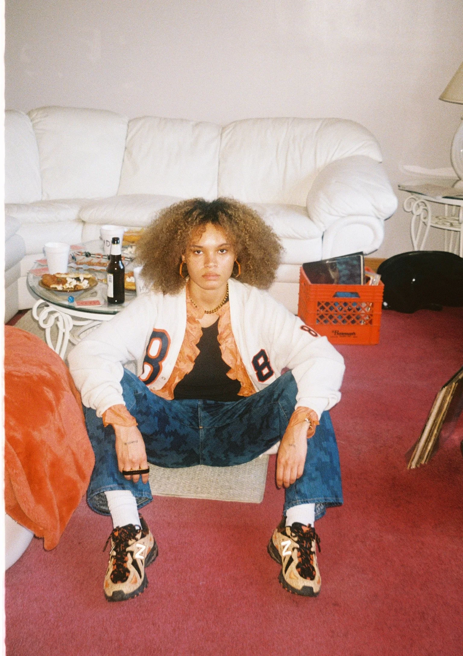 A young woman with curly hair sitting on the floor in a room with a red carpet, white couch, and a side table with drinks and snacks, wearing a white letterman jacket, dark shirt, camouflage jeans, and beige sneakers.