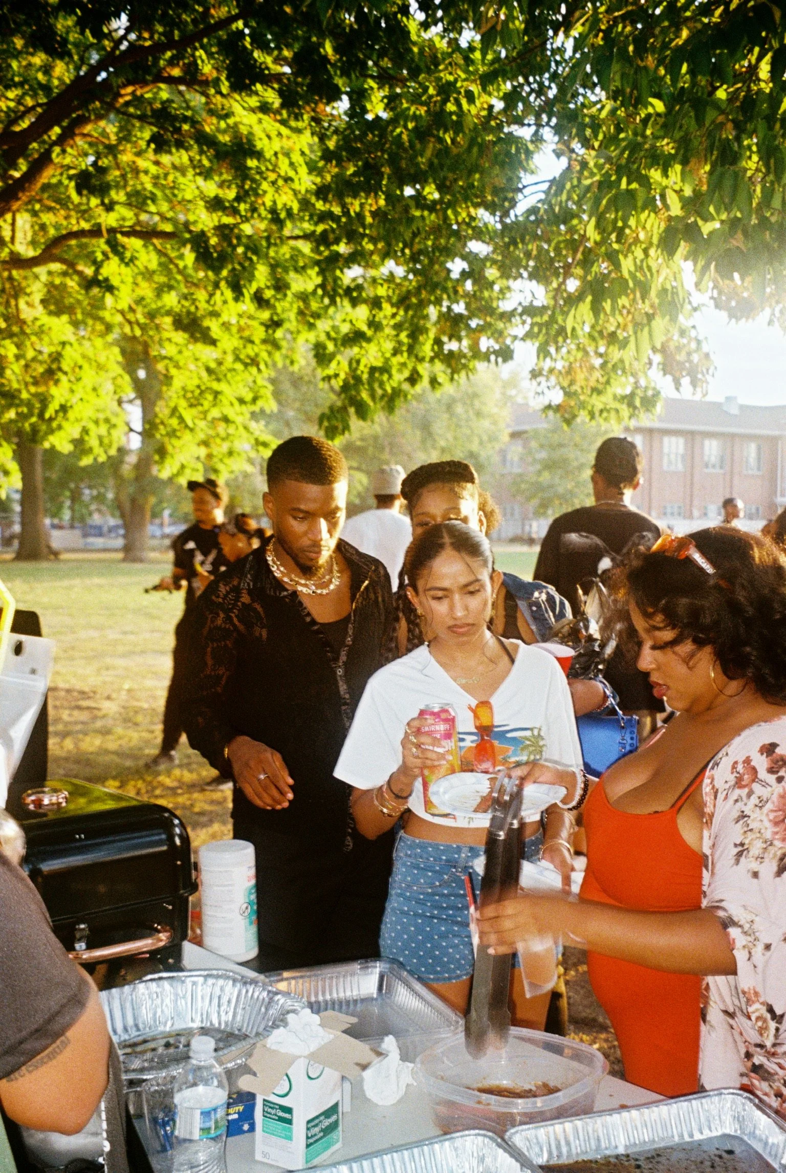 Group of diverse people gathered around a table at an outdoor barbecue in a park during daytime, with trees and buildings in the background.