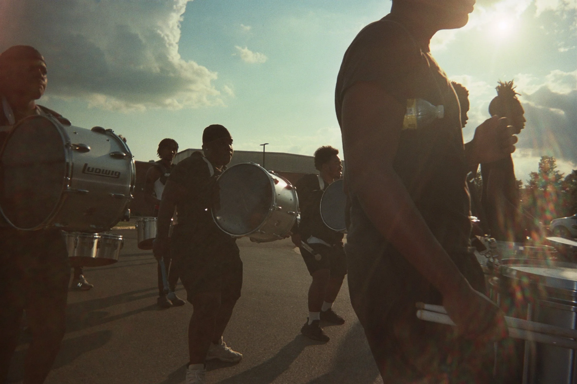 A group of musicians carrying drums and percussion instruments outside, with the sun shining behind them and a partly cloudy sky.