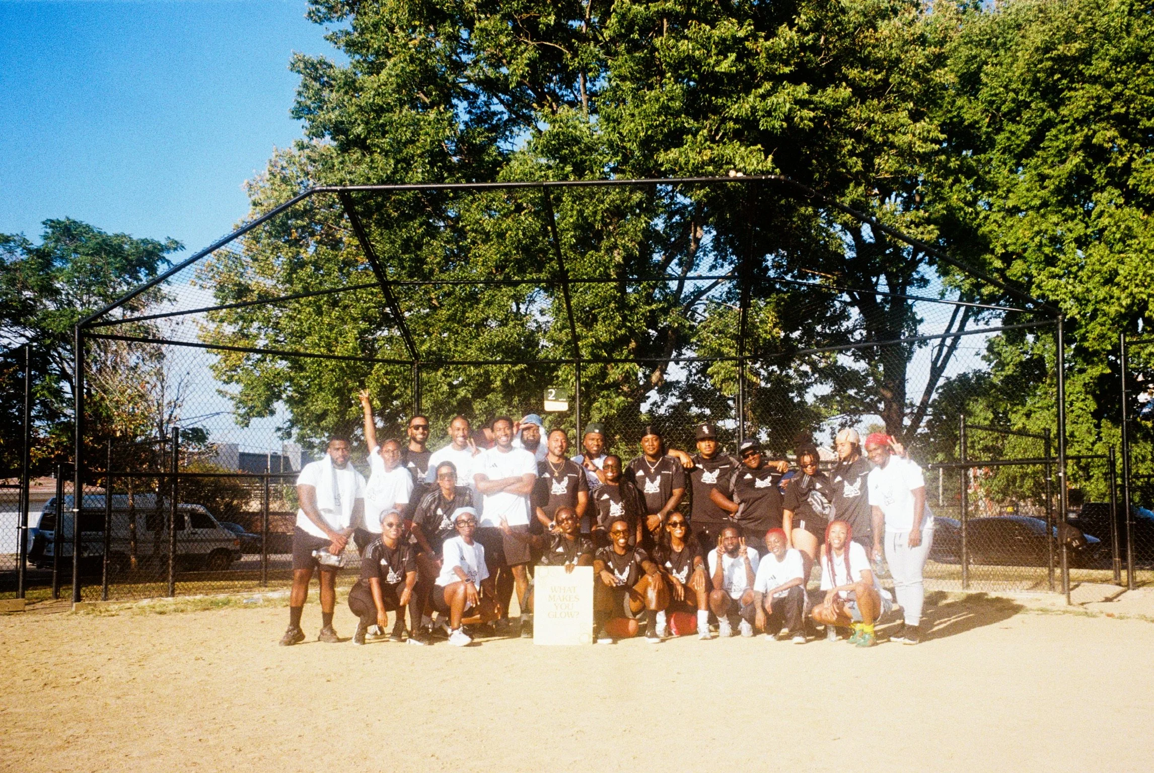 Group of people standing on a baseball field in front of a chain-link backstop, surrounded by green trees on a bright, sunny day.