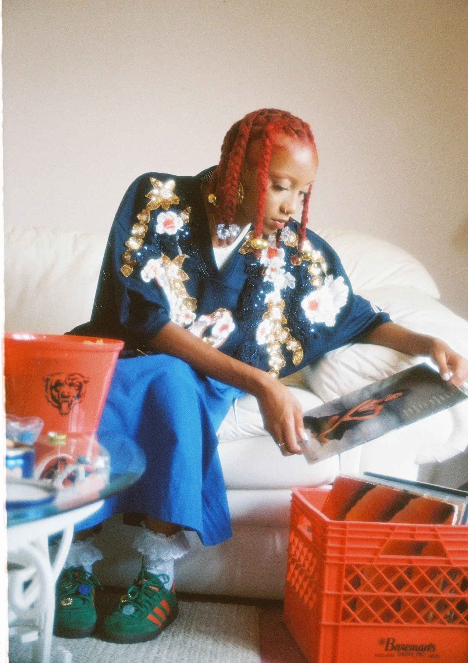 A woman with red braided hair is sitting on a white sofa, reading a magazine. She is wearing a dark blue jacket with floral, gold, and beaded embellishments, blue pants, white lace socks, and green Adidas sneakers with orange stripes. There is an orange bucket with a tiger logo and some other items on a small table nearby, and an orange crate on the floor filled with more magazines.