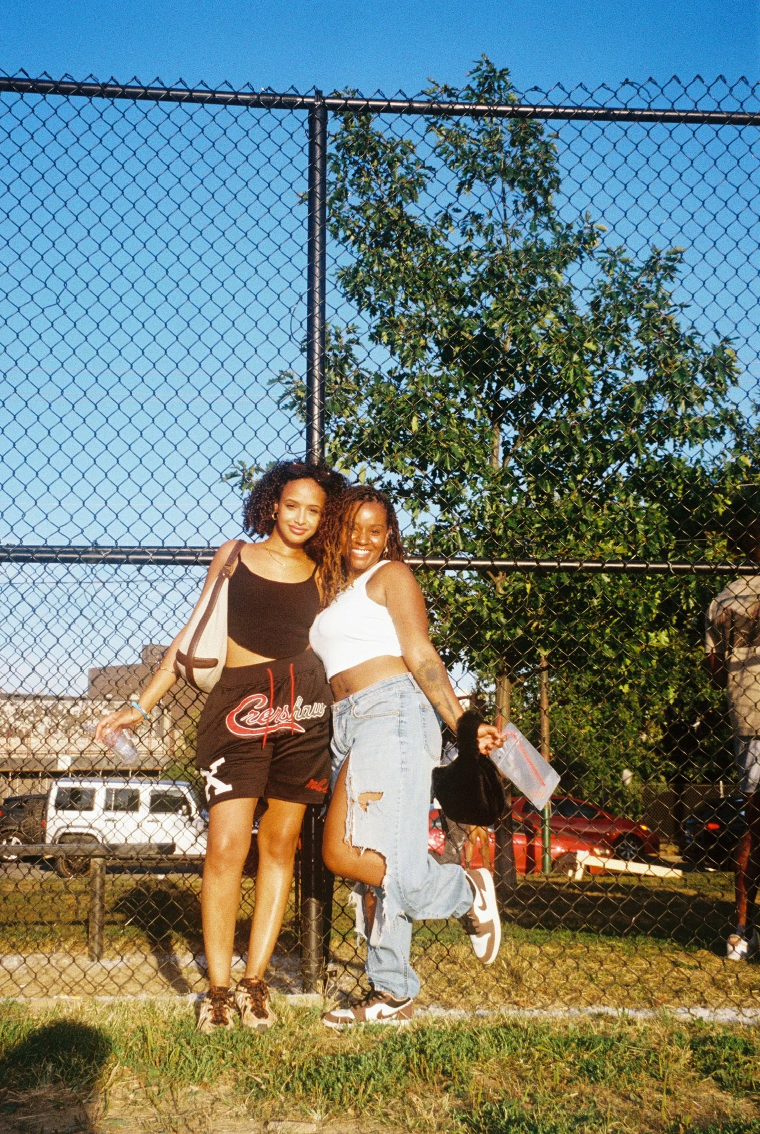 Two young women standing together in front of a chain-link fence on a sunny day, smiling for the camera, with a tree and parked cars visible in the background.