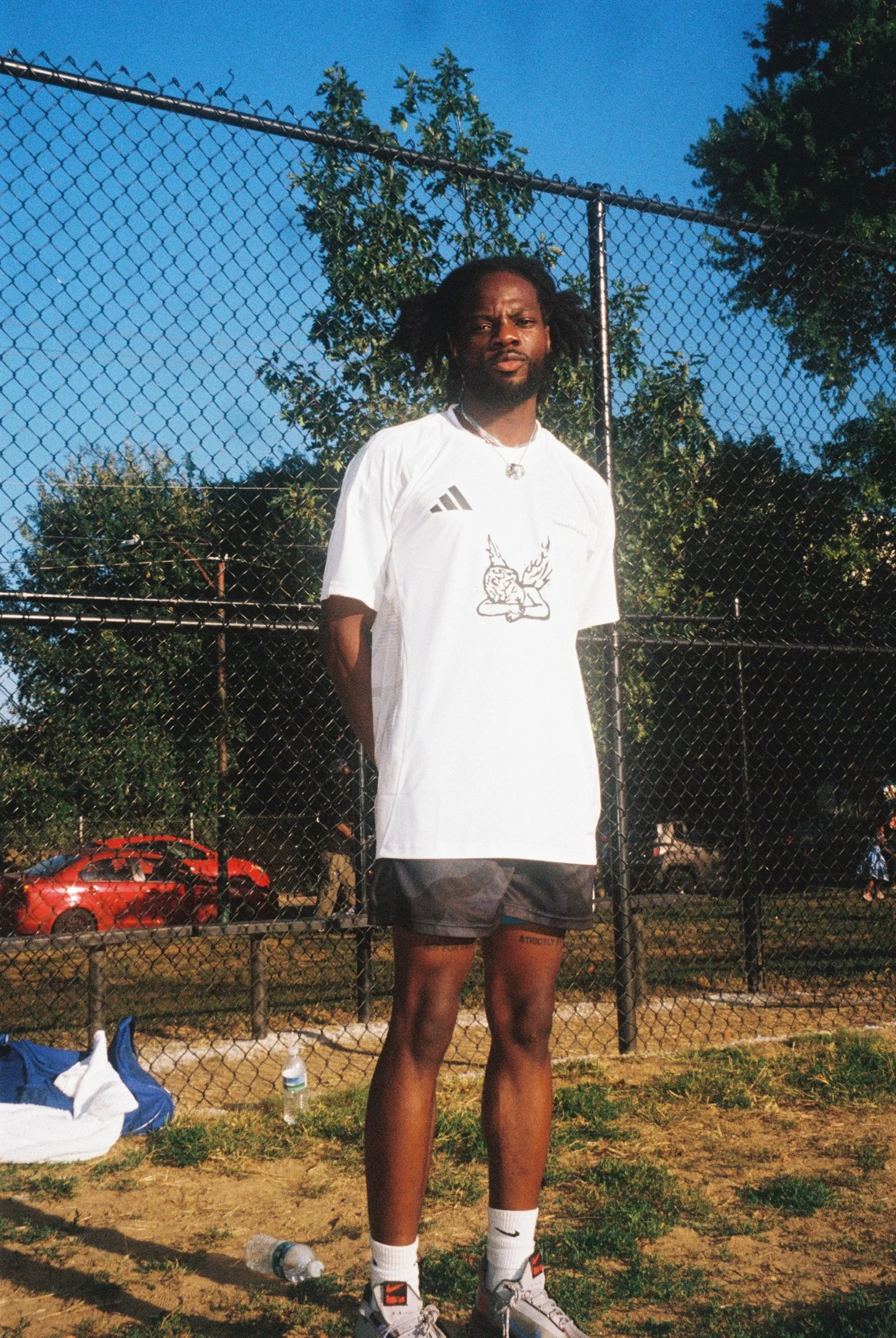A man stands outdoors in front of a chain-link fence, wearing a white sports jersey and black shorts, with trees and a red car in the background under a clear blue sky.