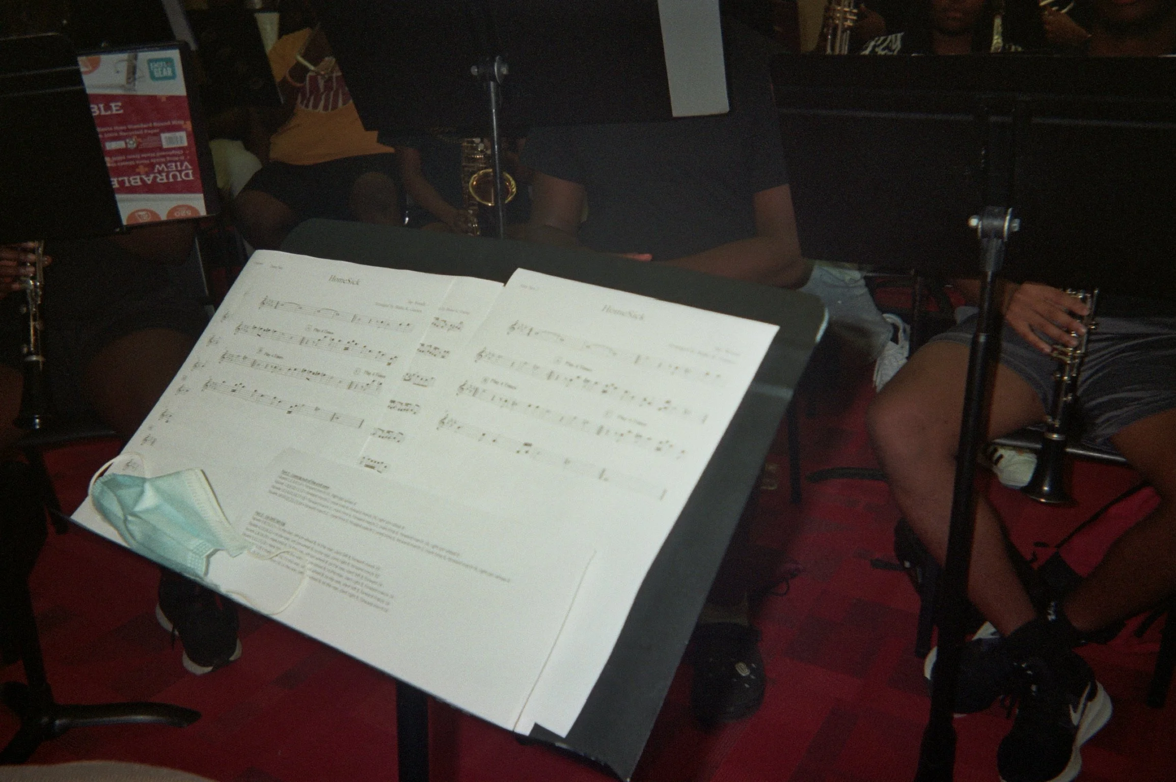 Music stand with sheet music in a music rehearsal room, with musicians holding flutes in the background.
