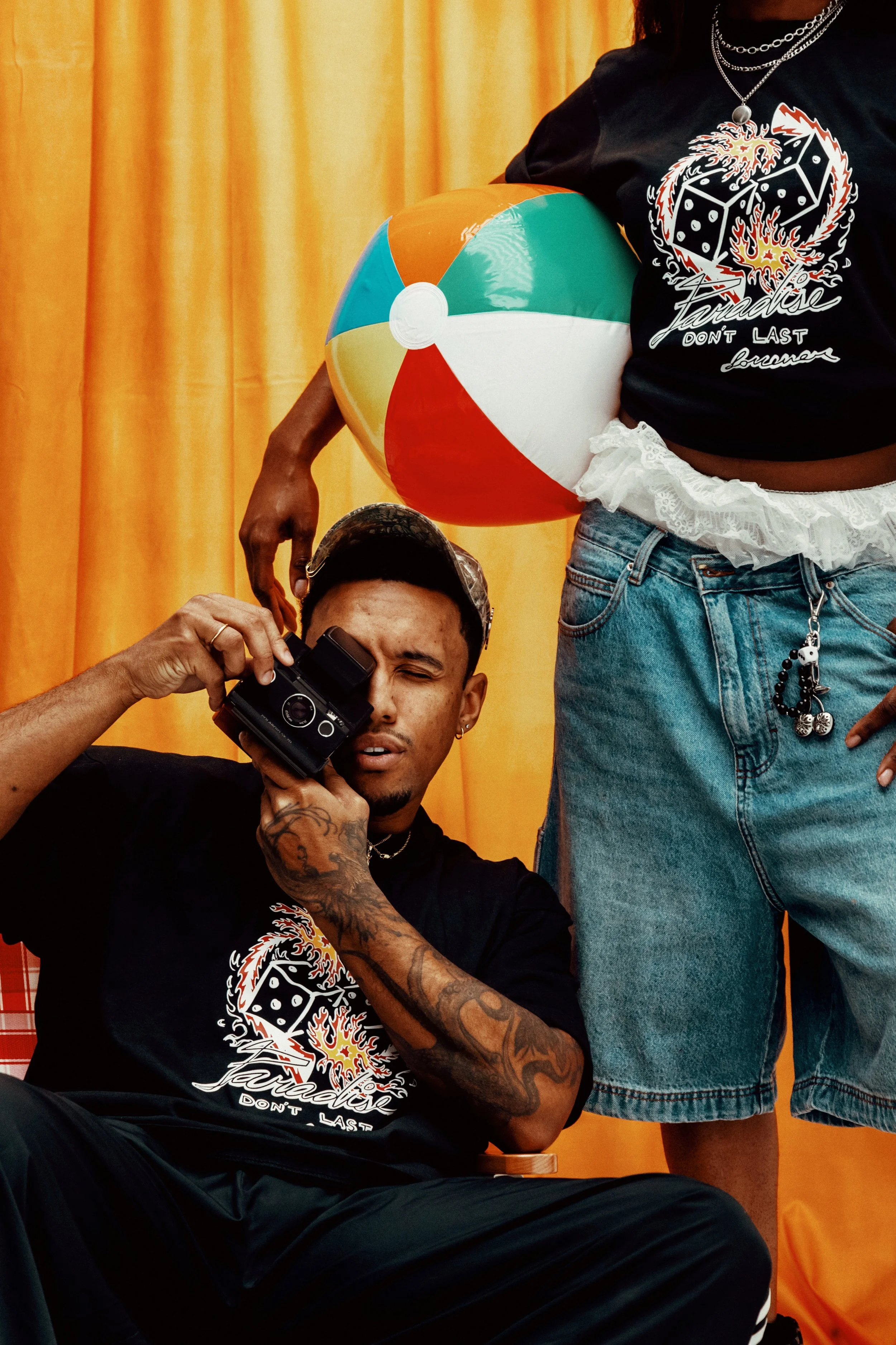 A man with tattoos on his arms is holding a camera to his eye and appears to be taking a photo. He is sitting in front of a yellow curtain and wearing a black t-shirt with a graphic design. Another person, wearing a black shirt and denim shorts, is standing next to him, holding a colorful beach ball.