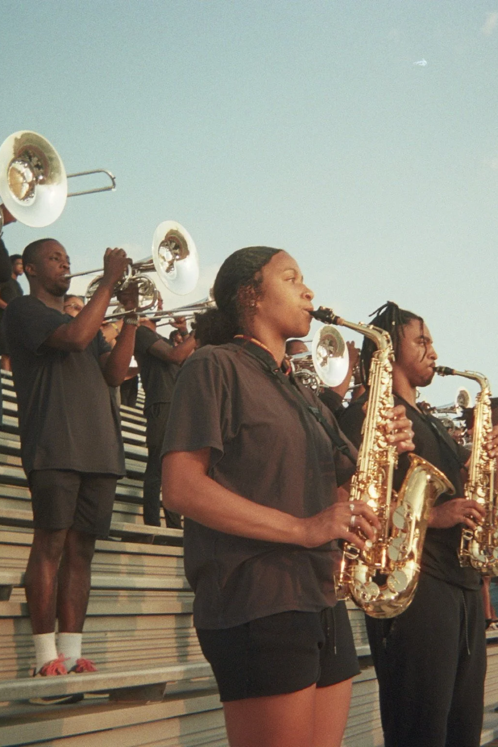 A group of musicians playing saxophones and trumpets outdoors, standing on bleachers against a blue sky.