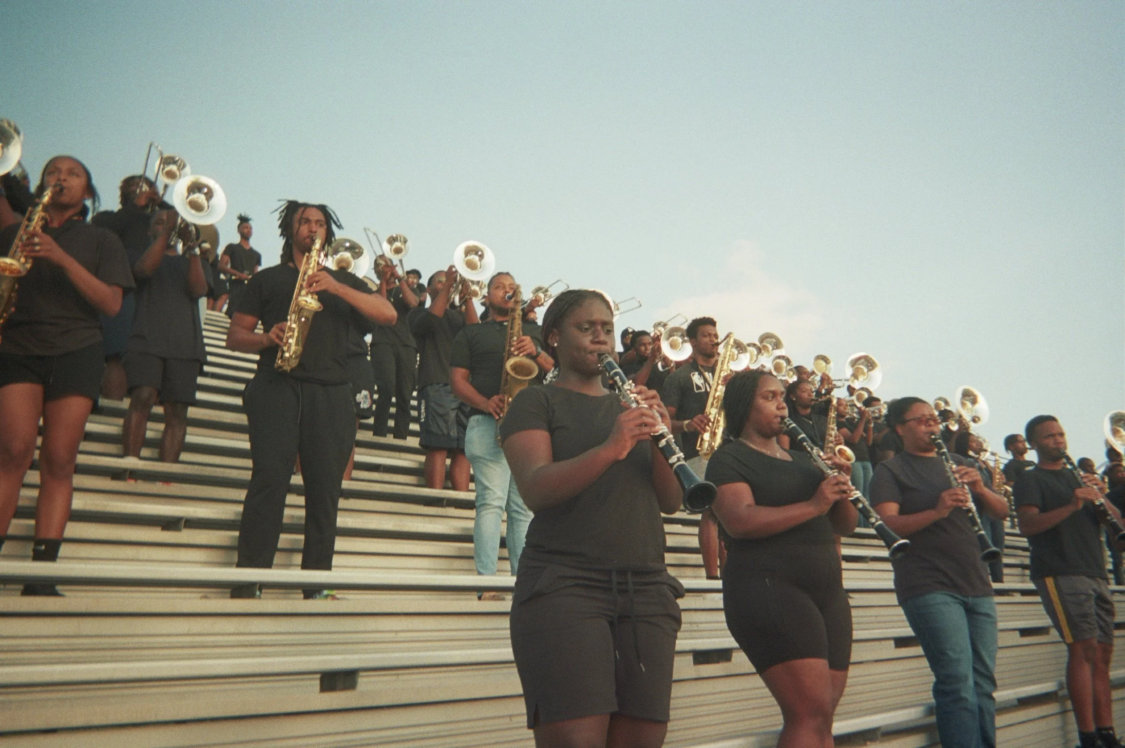 Group of musicians, mostly women, playing wind instruments such as clarinets and saxophones on outdoor bleachers.