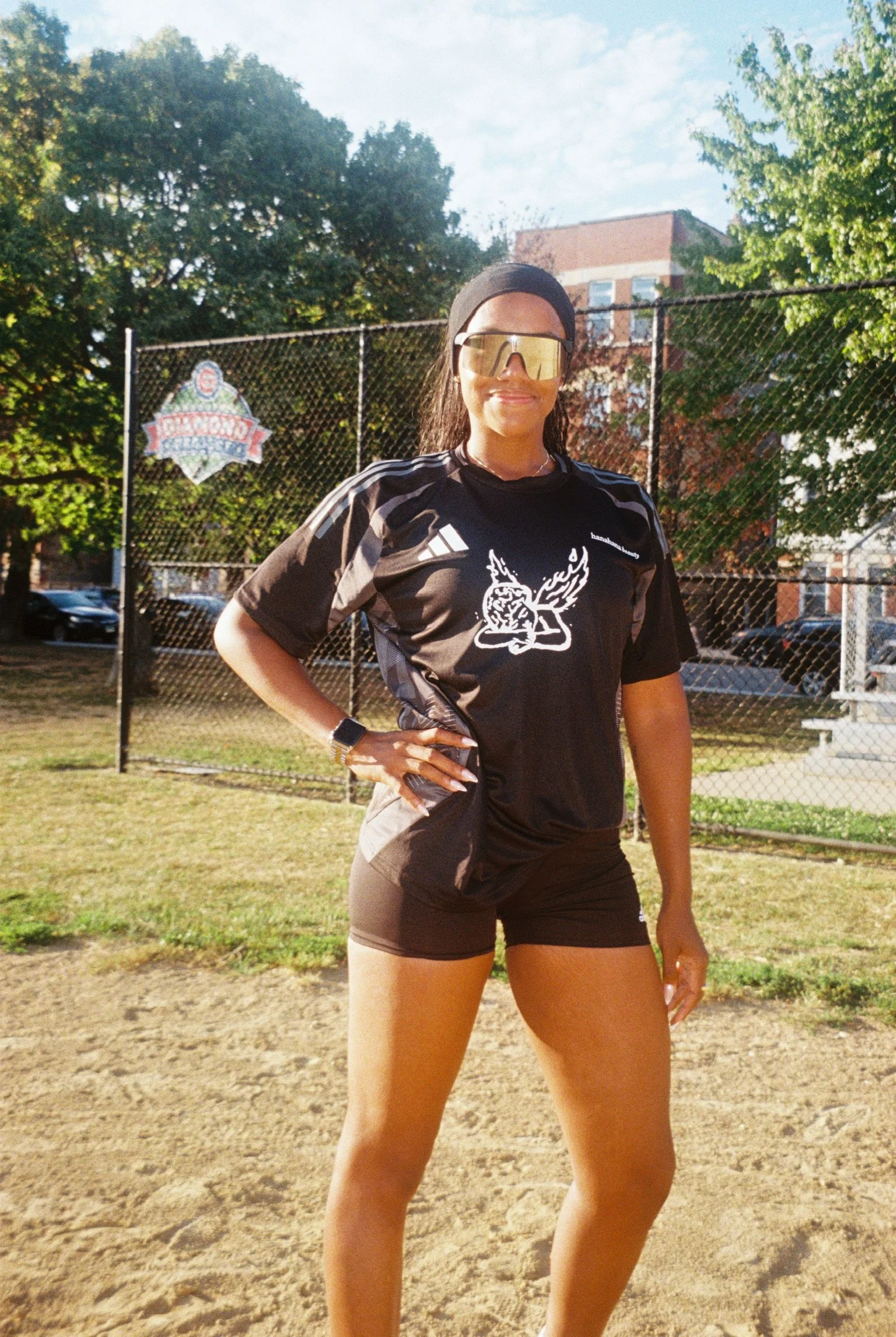 Woman in black athletic shirt and shorts standing outdoors with hand on hip, wearing large mirrored sunglasses and a black headband, on a sunny day in front of a chain-link fence.