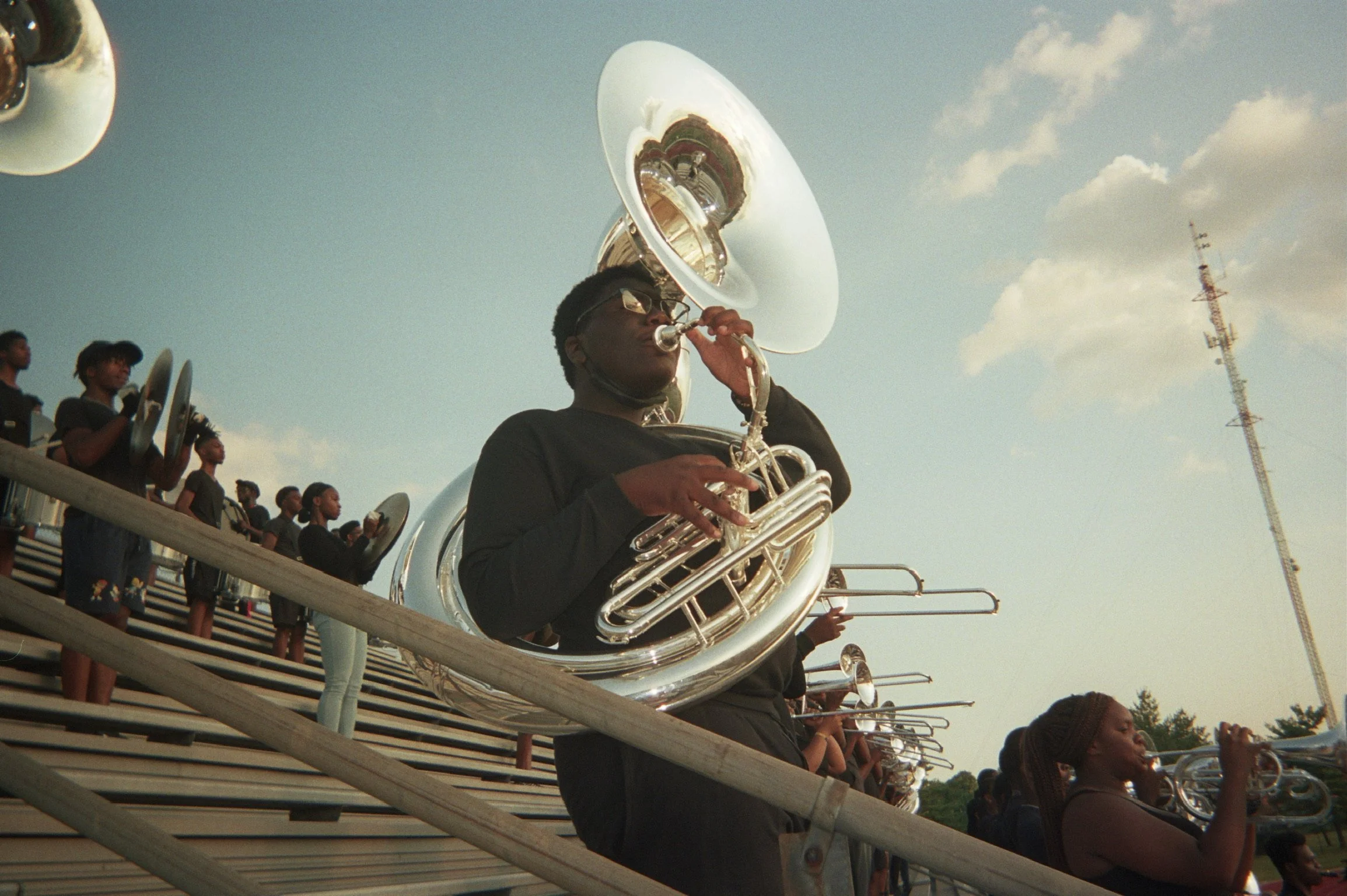 A marching band performing outdoors on bleachers, with a young man playing a sousaphone in the foreground and other band members playing brass instruments in the background.