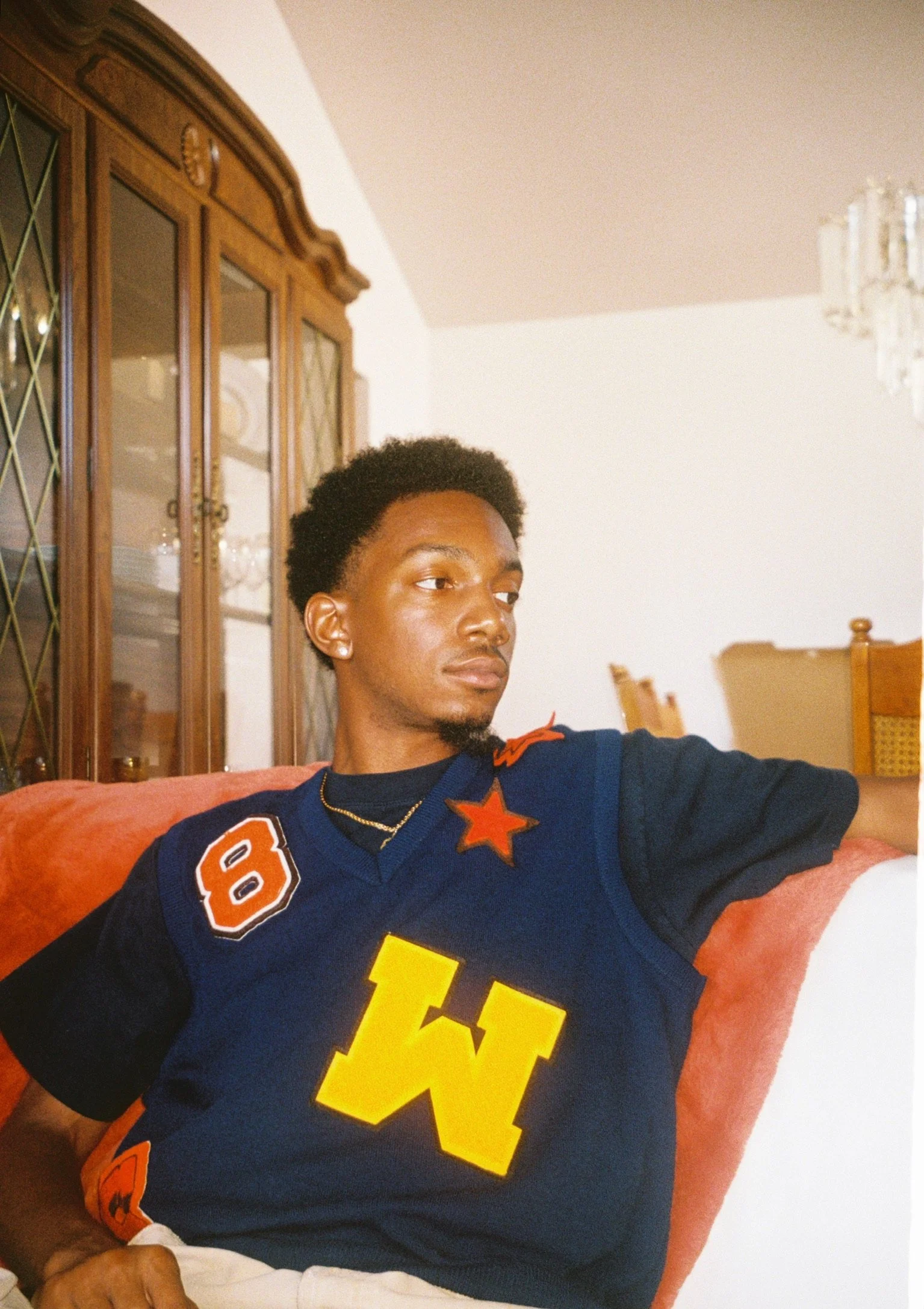Young man sitting on a couch wearing a navy blue sports jersey with orange and yellow patches and a large yellow letter 'W' on the front, in a living room with wooden china cabinet and chandelier.