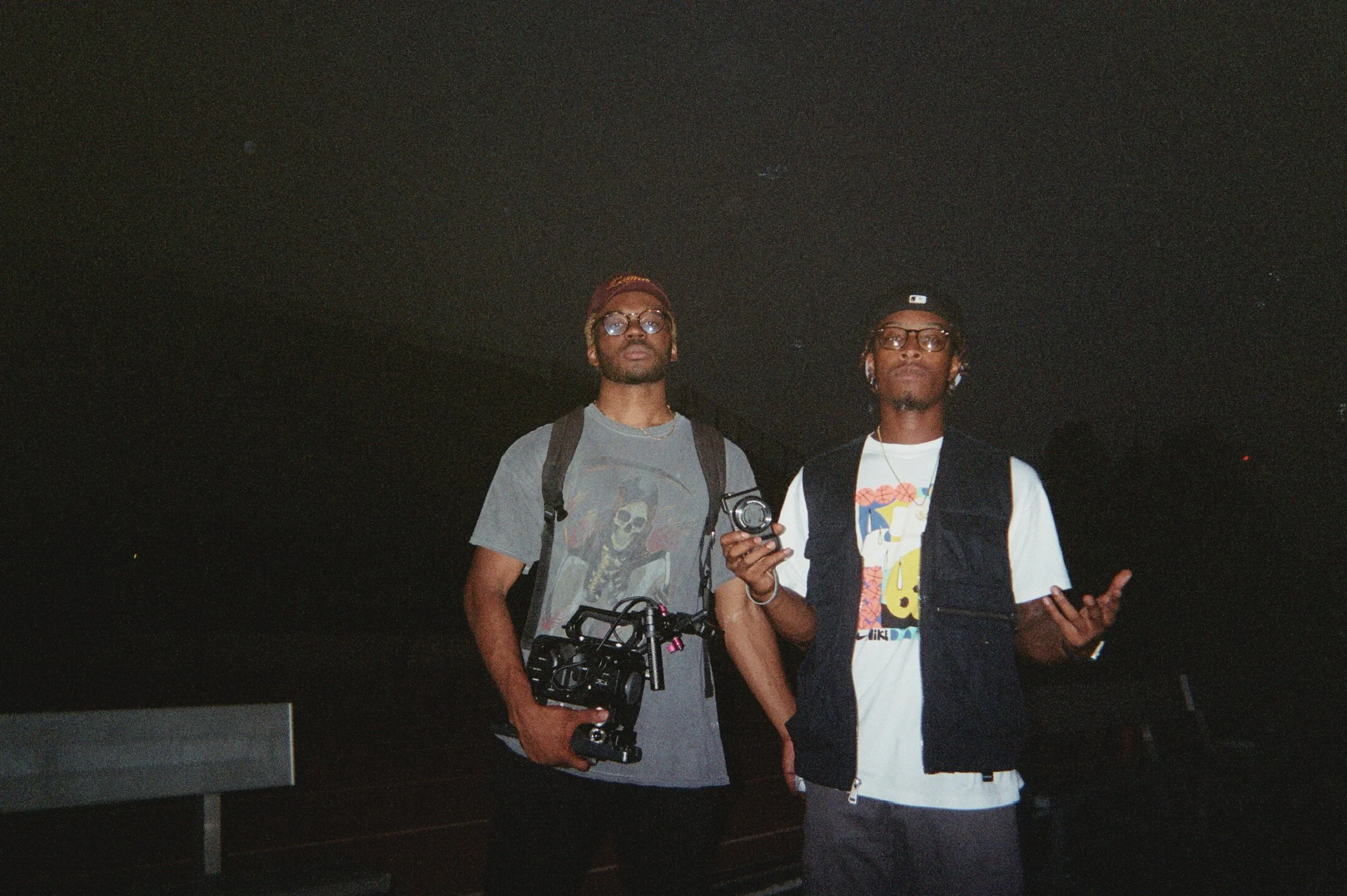 Two young men standing in a dark outdoor setting at night, both wearing glasses and casual clothing, with one holding a drone and the other holding a camera.