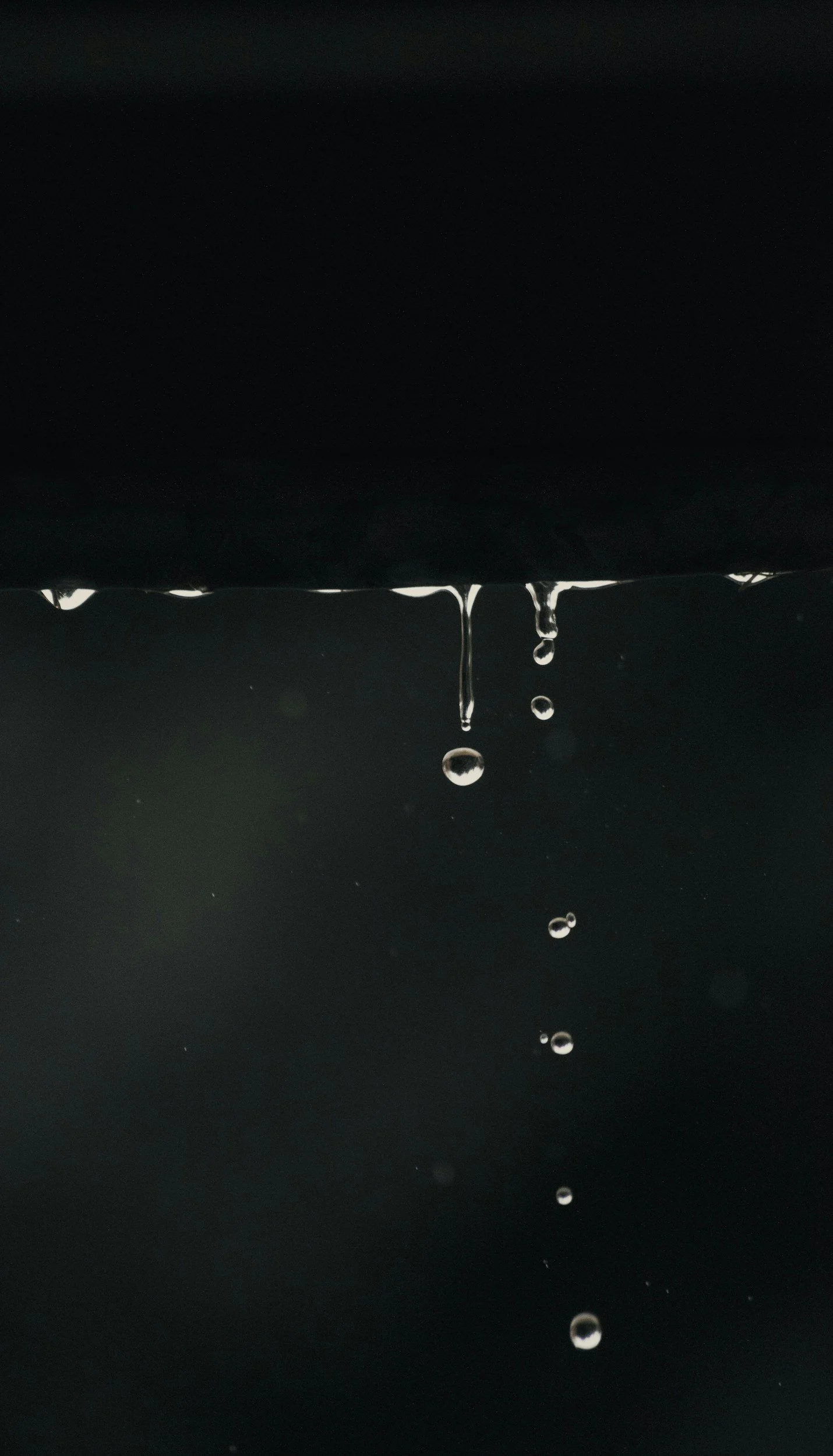 Close-up of water droplets falling from a dark surface against a dark background.