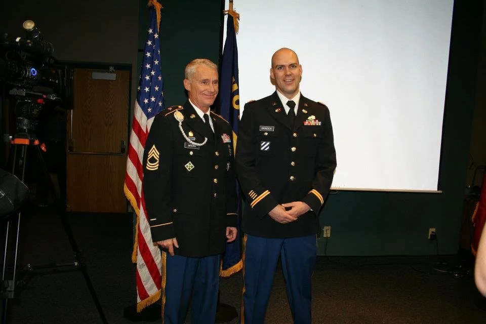 Two men in military uniforms standing side by side, smiling, with flags in the background and a presentation screen behind them.