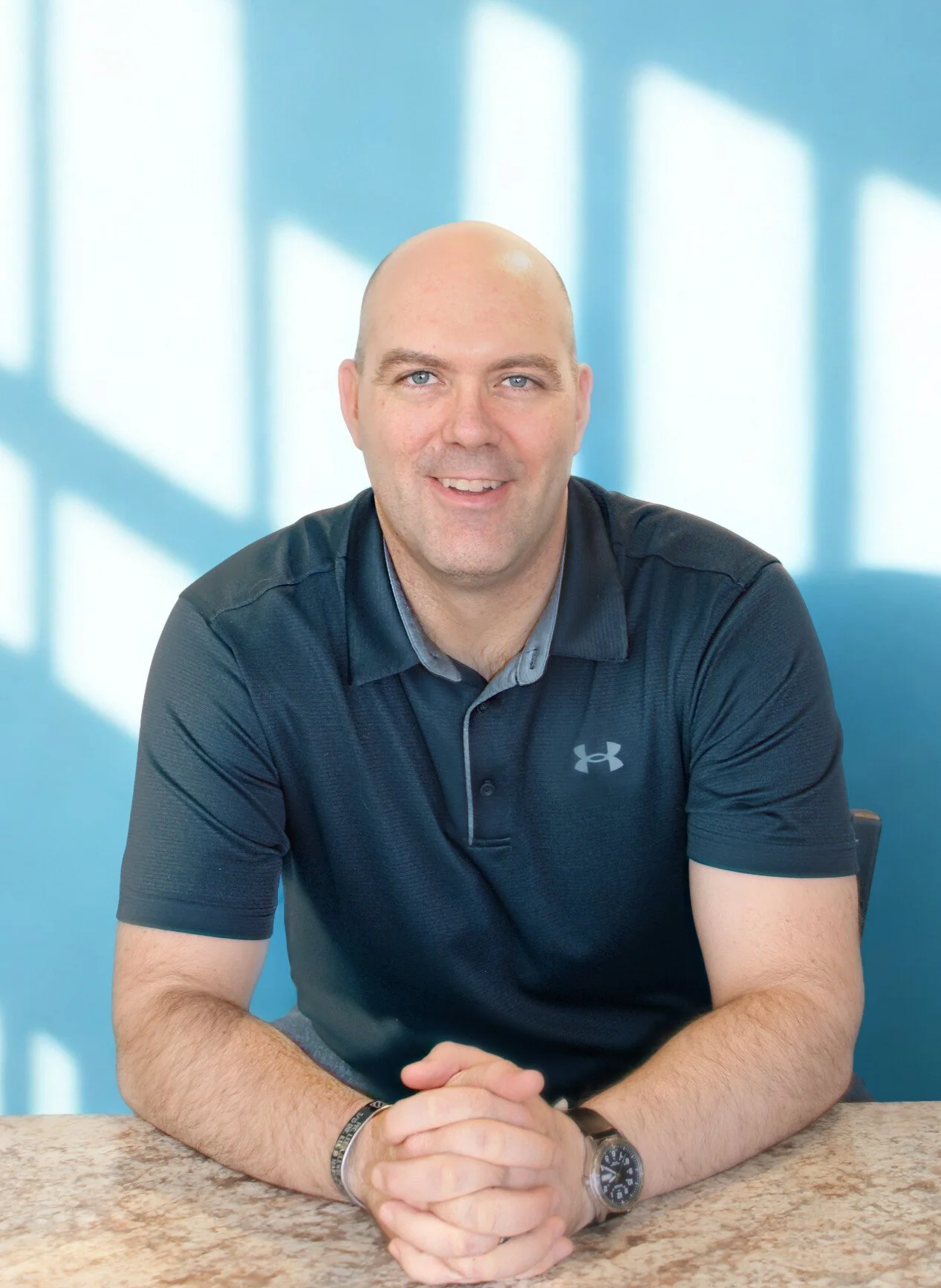 A smiling man with a shaved head, wearing a navy blue Under Armour polo shirt, sitting at a table with hands clasped, in front of a light blue wall with shadow patterns.