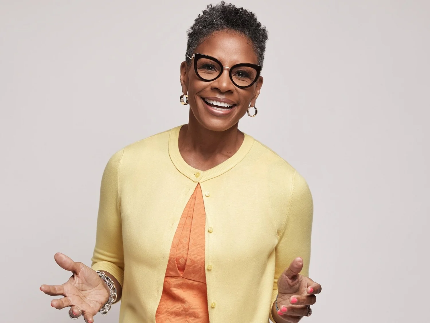 A smiling middle-aged woman with short curly gray hair wearing black glasses, gold hoop earrings, a yellow cardigan, and an orange top, standing against a plain light background.