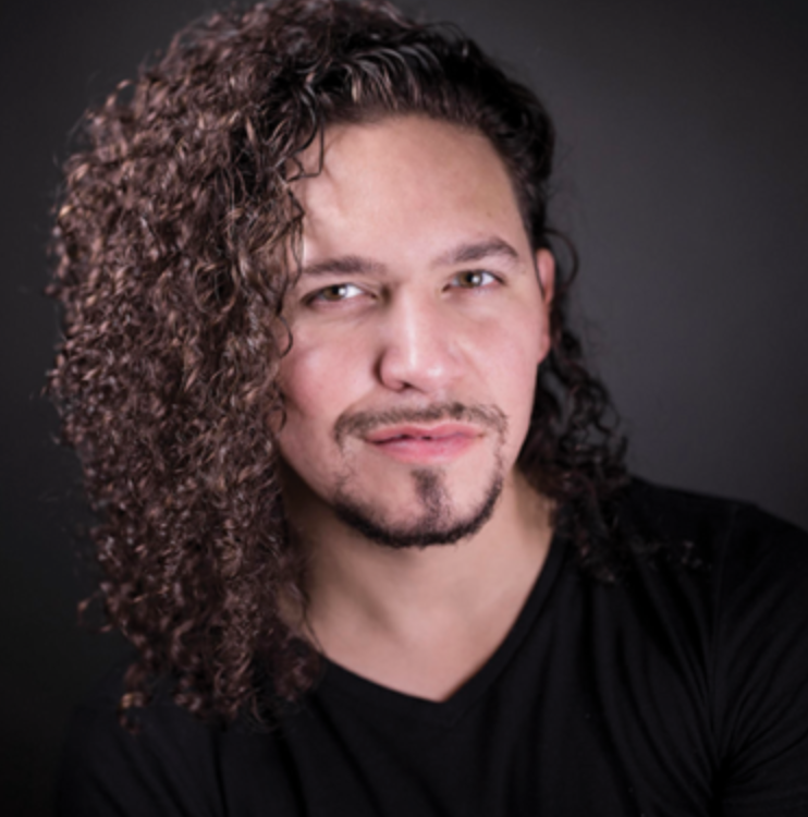 Portrait of a man with curly brown hair and facial hair, wearing a black shirt, against a dark background.