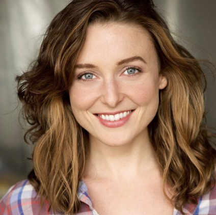 Close-up portrait of a young woman with wavy, shoulder-length brown hair, blue eyes, and a bright smile, wearing a plaid shirt.