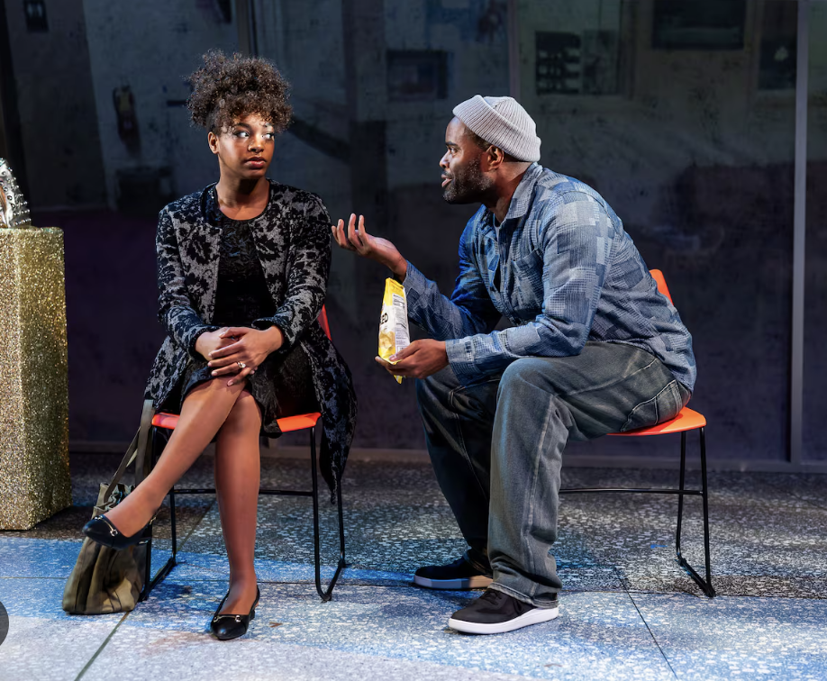 A man and woman sitting and talking, the woman looks uninterested while the man is gesturing with his hands, holding a bag of chips.