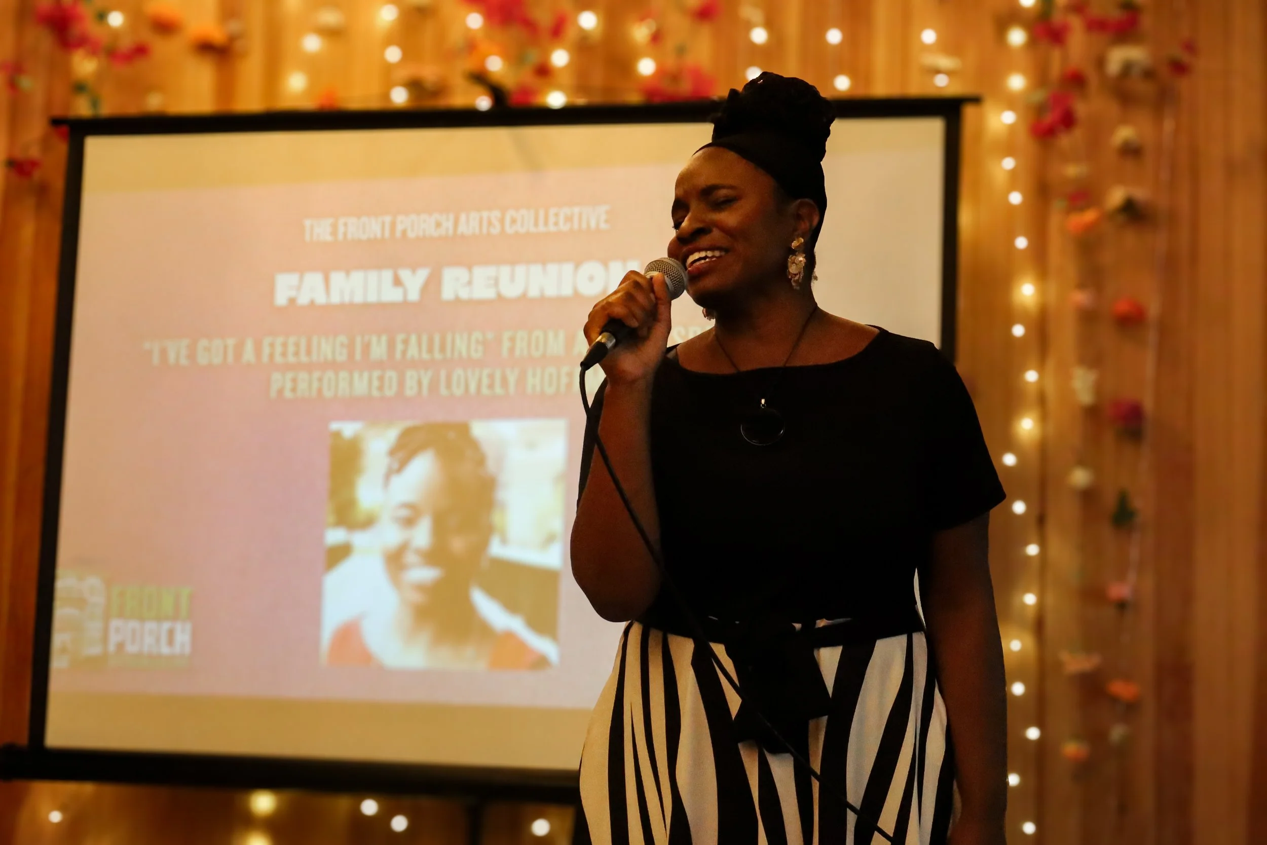 A woman speaking into a microphone at a family reunion event, standing in front of a large screen displaying the event details, with warm decorative lighting in the background.