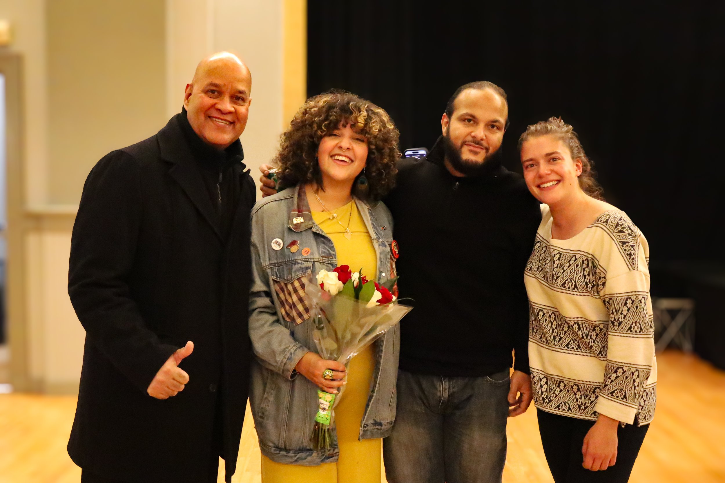 Four people standing together smiling, one woman holding a bouquet of flowers. Two men and two women, all in casual or semi-formal attire, inside a room with warm lighting.