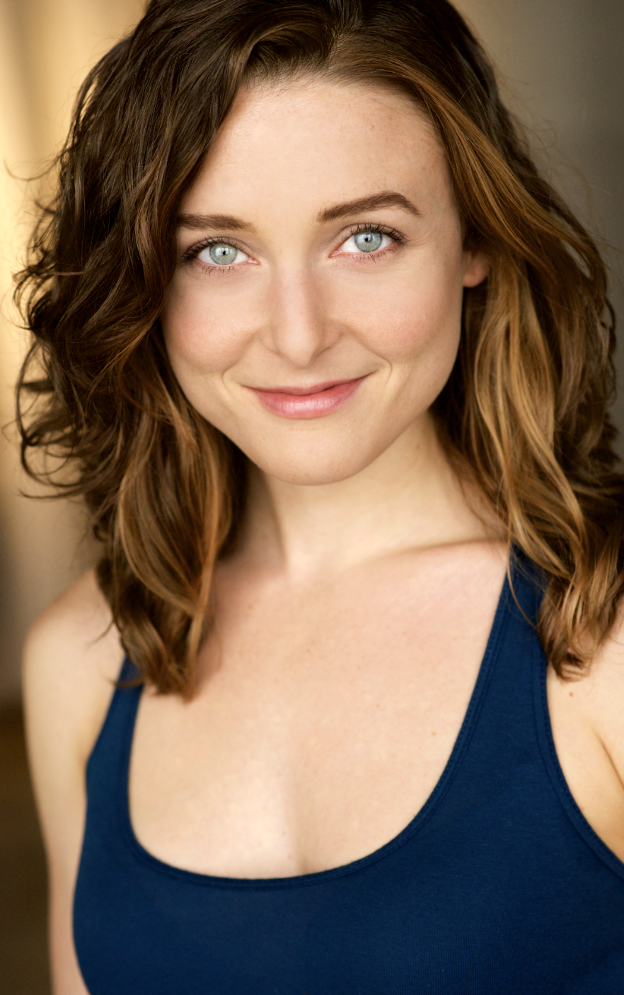 A woman with shoulder-length curly brown hair and blue eyes, smiling and wearing a blue sleeveless top.