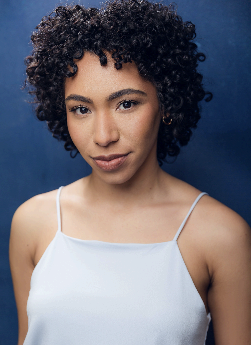 A young woman with curly black hair and light brown skin, wearing a white spaghetti strap top, standing against a dark blue background.