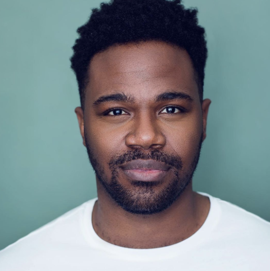 Close-up portrait of a young Black man with short curly hair and facial hair, wearing a white T-shirt, against a muted green background.