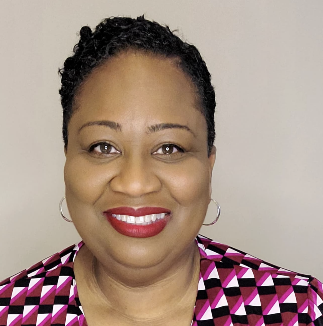 Close-up portrait of a smiling woman with short curly hair, red lipstick, hoop earrings, and a patterned blouse against a plain background.