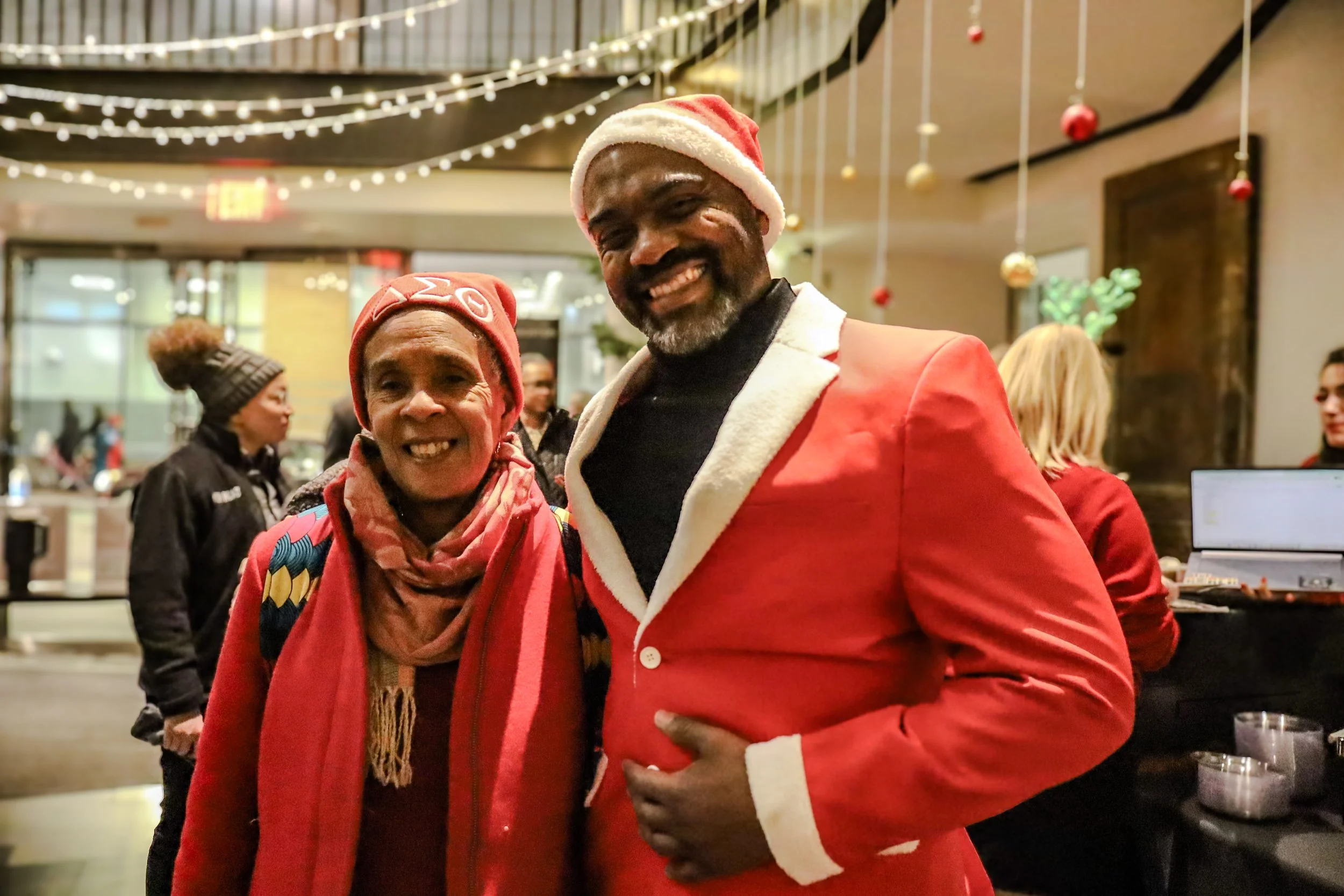 A smiling man dressed as Santa Claus and an older woman smiling, both wearing Christmas-themed hats, at a festive indoor holiday event with decorations and string lights.