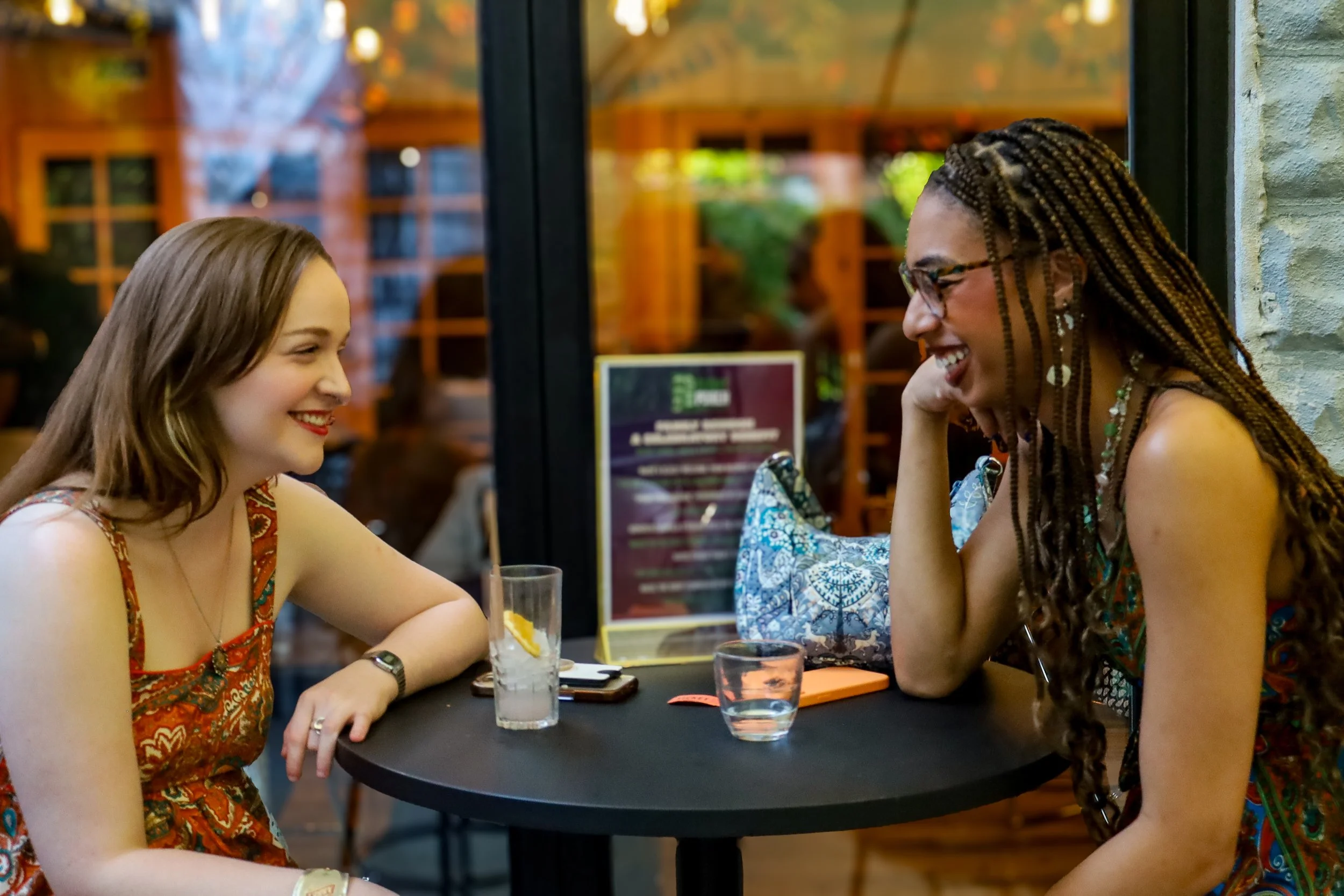 Two women sitting at a black table in a cozy indoor setting, smiling and engaging in conversation. One woman has long blonde hair and the other has long braided hair, both wearing colorful dresses. There are drinks on the table and a blurred backgrou