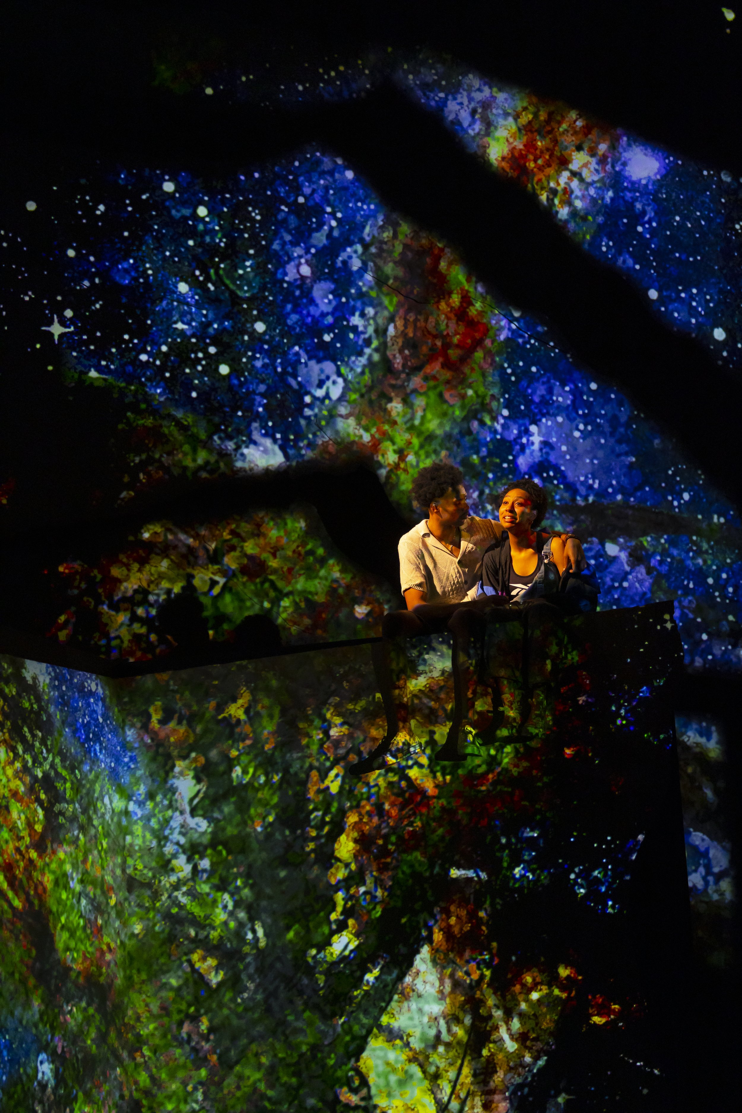 Two women sitting on a black wrought iron bench, surrounded by a vibrant, colorful, starry digital art background. One woman with curly hair is wearing a white top, and the other with short hair is wearing a black jacket and striped shirt. They are close to each other, smiling and talking.