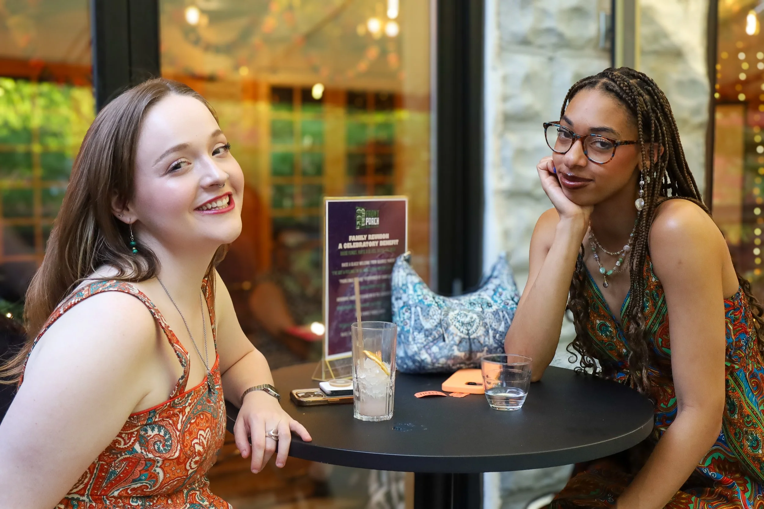 Two women with long hair and glasses sitting at a table, smiling and resting their heads on their hands in a restaurant.