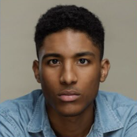 Portrait of a young man with short curly hair, wearing a light blue denim shirt, looking directly at the camera against a plain background.