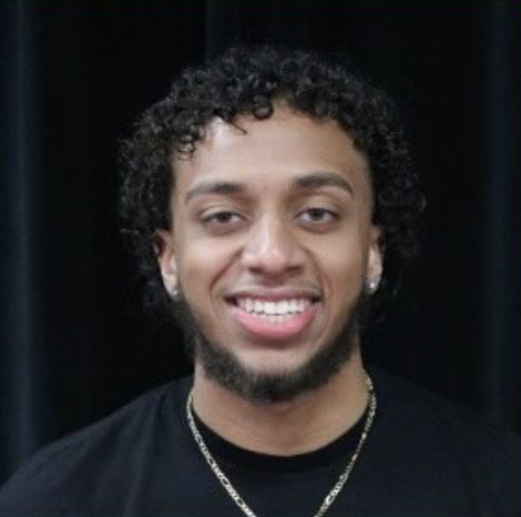 A young man smiling with curly hair, wearing earrings, a chain necklace, and a black shirt, against a dark background.