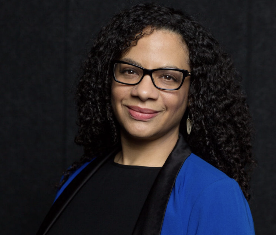 A woman with curly hair, wearing glasses and a blue blazer, smiling in front of a dark background.