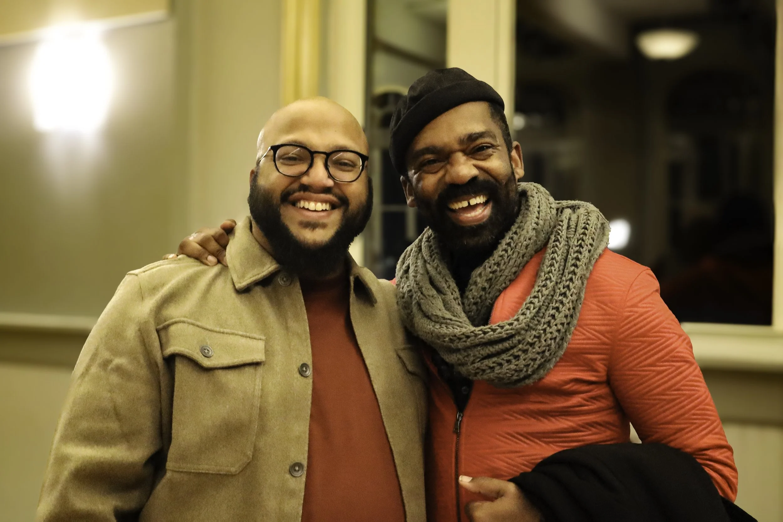 Two men smiling and standing close together indoors. One is wearing glasses, a tan jacket, and an orange shirt; the other has a beard, is wearing a black hat, a scarf, and a red jacket.