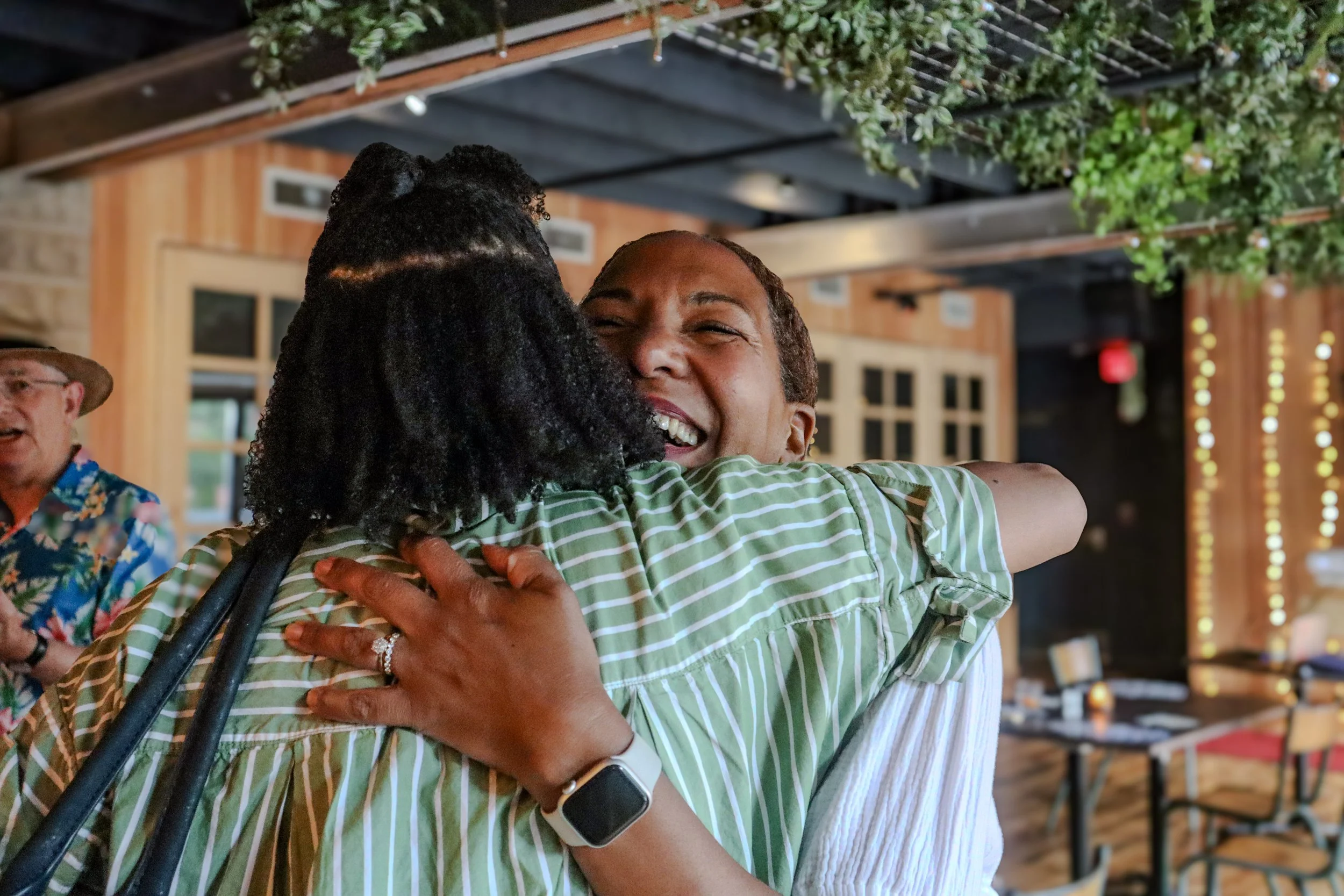 Two women hugging each other and smiling in a lively indoor setting with wooden walls, ceiling beams, and string lights.