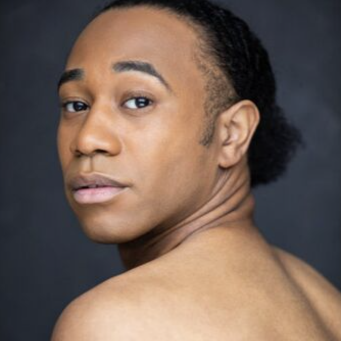 Close-up portrait of a young man with dark hair styled in a ponytail, looking over his shoulder at the camera against a dark background.