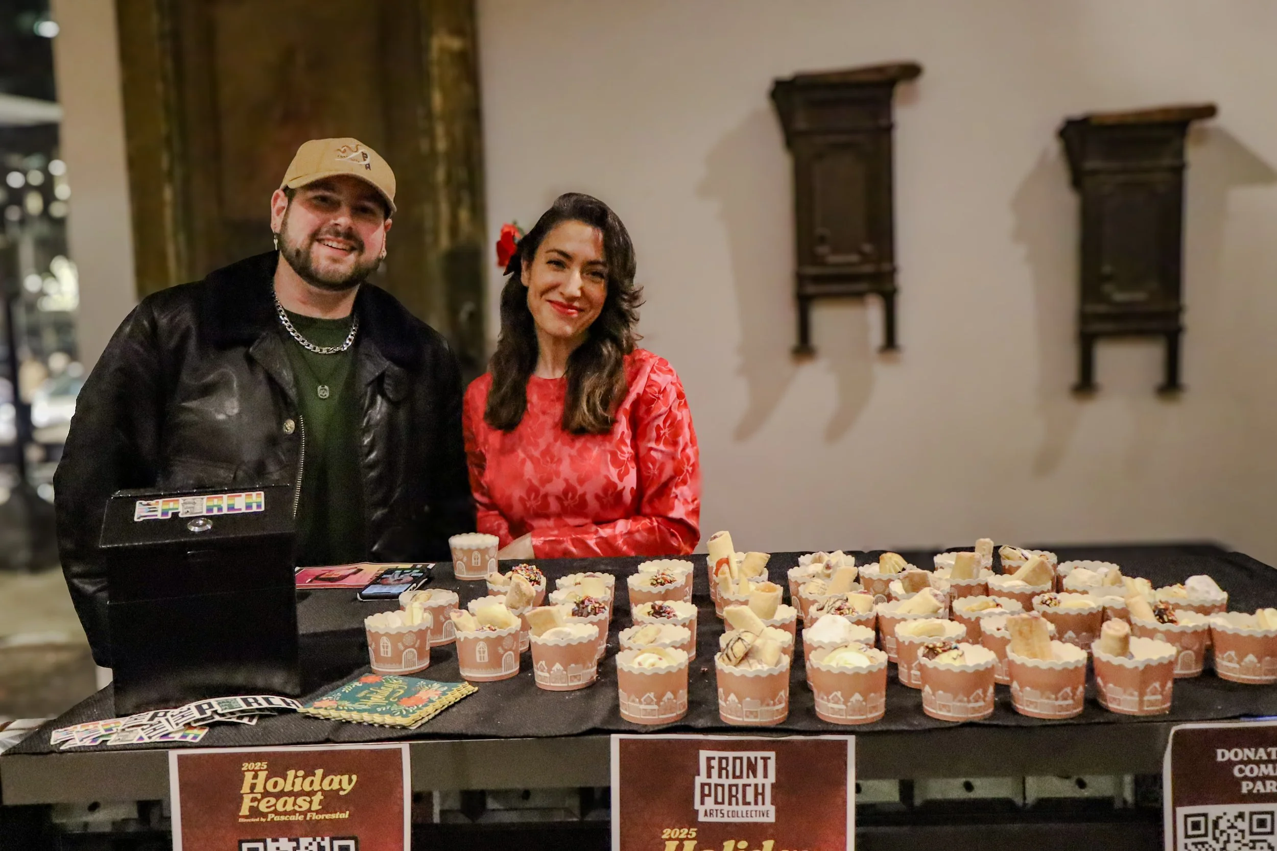 A man and woman standing behind a table with decorated cupcakes at a holiday event. The man is wearing a tan cap, black leather jacket, and silver chain, and the woman is wearing a red dress with floral pattern and has a red flower in her hair. The t