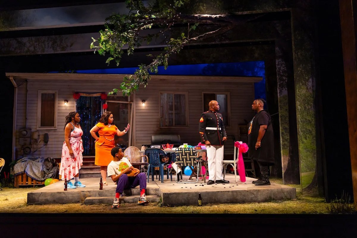 The photo shows a group of six people on a backyard stage, with a house in the background. Two men are in animated conversation in the center, one in a military uniform, the other in a black hoodie. Three women and one young boy are gathered around, with the women expressing different emotions. The scene appears to be from a theatrical play, with props, colorful balloons, and outdoor furniture.
