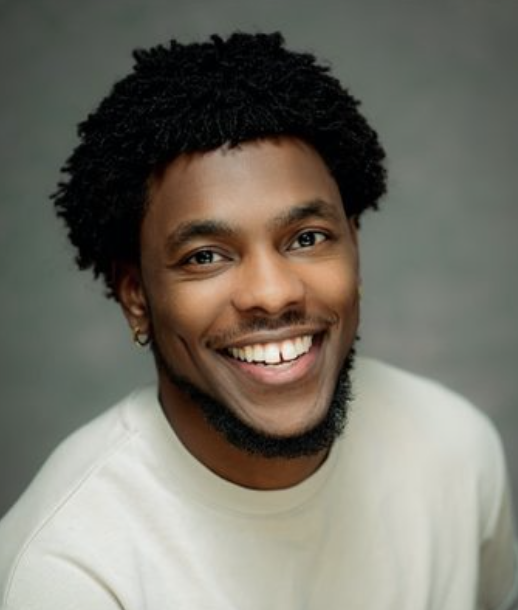 Smiling African American man with curly hair and beard, wearing a white shirt against a gray background.