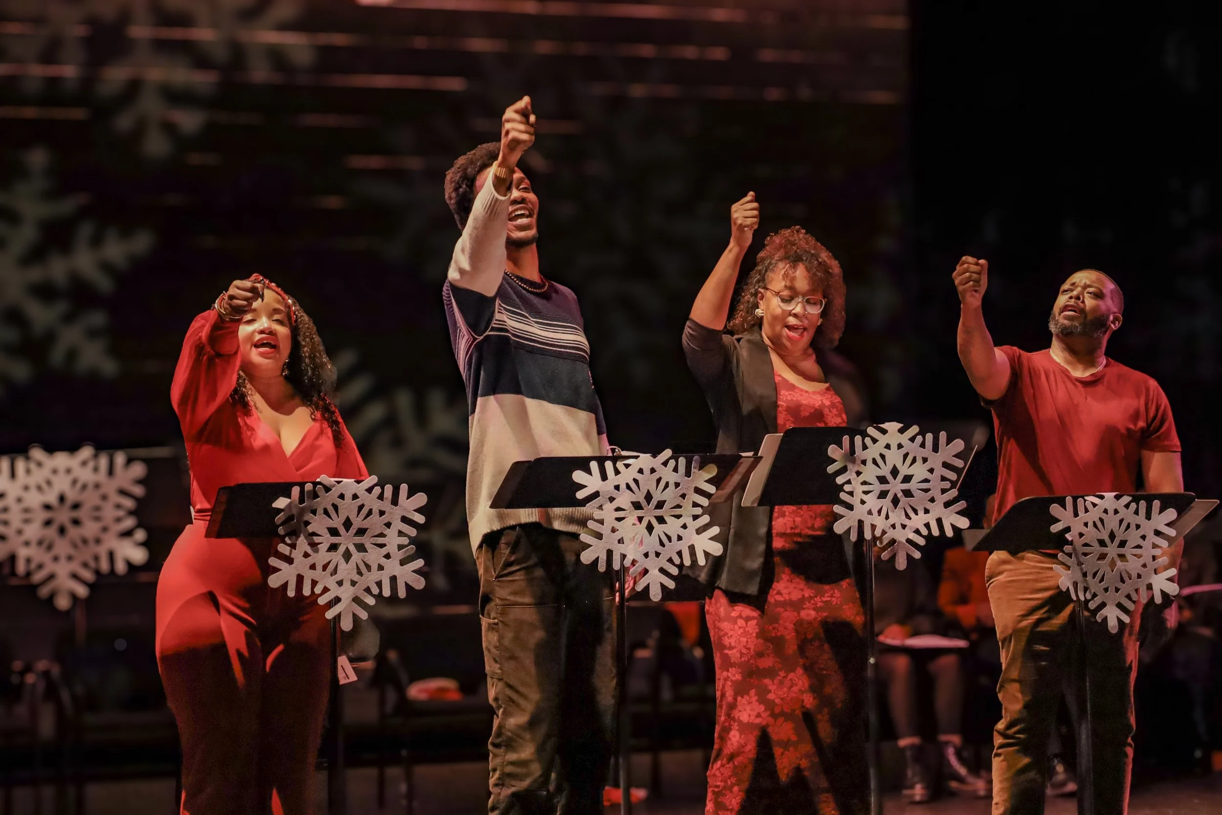 Four people singing or performing in a group on stage, with Christmas snowflake decorations on music stands, during a holiday concert.