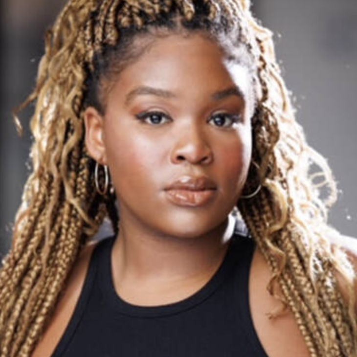 A woman with blonde dreadlocks wearing hoop earrings and a black sleeveless top, looking directly at the camera.