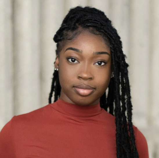 Portrait of a young African American woman with long braided hair wearing a red turtleneck sweater against a neutral background.